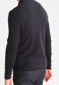 Man som bär en mörkgrå långärmad tröja och svarta byxor, visad bakifrån mot en vit bakgrund.