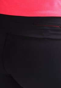 Lähellä otettu kuva mustista urheilulegginsien vyötäröstä, jossa on pieni vetoketjullinen tasku, yhdistettynä kirkkaanpunaiseen ribbitoppiin.