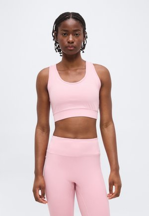 Νεαρή γυναίκα με πλεξούδες, που φοράει ροζ αθλητικό σουτιέν και κολάν με υψηλή μέση, στέκεται μπροστά σε απλό ανοιχτό φόντο.