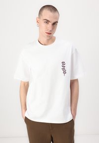White cotton T-shirt featuring a small dark floral design on the left chest. Loose fit, round neckline, and short sleeves.