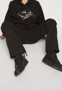 Czarna bluza z kapturem z grafiką kasety, w zestawie z czarnymi spodniami typu cargo i czarnymi sneakersami Nike. Wszystkie elementy mają matowe wykończenie i luźny krój.