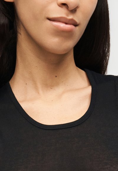 Το κάτω μέρος του προσώπου και ο λαιμός γυναίκας που φοράει μαύρο τοπ, εμφανίζοντας καθαρό δέρμα και φυσικά ροζ χείλη με ένα διακριτικό χαμόγελο.