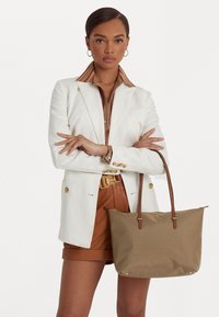 Saco tote castanho com alças de couro, textura de tela suave, detalhes em metal dourado e logotipo discreto. Usado com um blazer branco e calções castanhos.