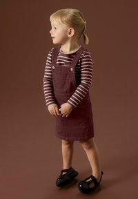 Une jeune enfant avec des cheveux blonds en queue de cheval, portant un t-shirt à rayures à manches longues, une robe salopette bordeaux et des chaussures noires, se tenant sur un fond brun, regardant vers la gauche.