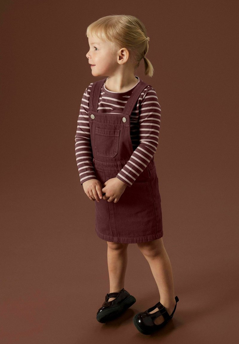 Une jeune enfant avec des cheveux blonds en queue de cheval, portant un t-shirt à rayures à manches longues, une robe salopette bordeaux et des chaussures noires, se tenant sur un fond brun, regardant vers la gauche.