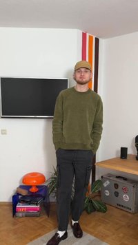 Olivgrön, texturerad tröja, grå cargobyxor, mörka skor. Bakgrunden innehåller en TV, orange lampa och trä möbler med dekorativa föremål.