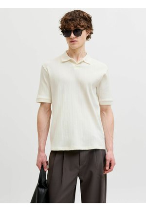 Jeune homme portant des lunettes de soleil noires, une chemise à manches courtes texturée couleur blanc cassé, un pantalon foncé, tenant un sac en cuir noir sur un fond uni.