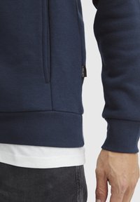 Felpa blu navy con polsini e orlo a coste, dotata di una tasca laterale e cuciture in vista. Indossata sopra una t-shirt bianca.