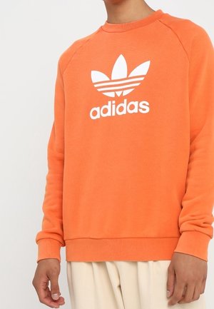 Person trägt einen orangefarbenen Adidas-Sweatshirt mit weißem Logo und Schriftzug, kombiniert mit hellbeigen Hosen und steht vor einem einfarbigen Hintergrund.