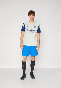 Nike football kit featuring a light gray top with dark blue sleeves, "Qatar Airways" logo, blue shorts, black socks, and black shoes.