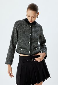 Giacca in tweed bianco e nero con design cropped, scollatura rotonda e grandi bottoni allacciati, abbinata a una gonna nera plissettata.