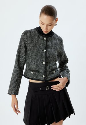 Veste en tweed noir et blanc avec un design court, un col rond et de grands boutons, associée à une jupe plissée noire.