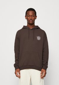 Hoodie marron avec une poche avant, capuche à cordon de serrage et logo soleil brodé sur la poitrine gauche. Texture de tissu douce, coupe décontractée.