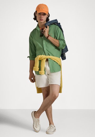 Grön skjorta med knappar, vita jeansshorts, gul tröja knuten i midjan, orange keps. Lätta sneakers fulländar looken.