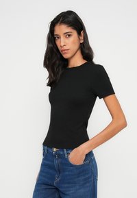 Μαύρο τοπ με κοντά μανίκια, εφαρμοστή γραμμή και λεία υφή, συνδυασμένο με μπλε ψηλόμεσα τζιν με τσέπες και ραφές.