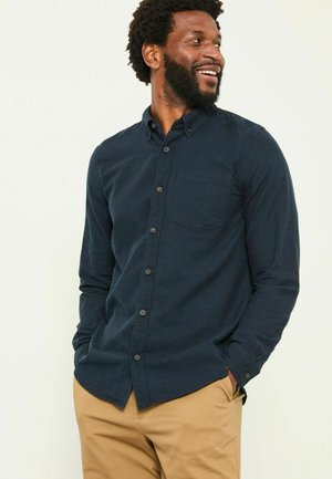 Navyblaues Langarmhemd mit Knopfverschluss, Brusttasche und leicht abgerundetem Saum. Getragen mit beigen Hosen, die einen strukturierten Stoff aufweisen.