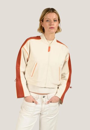 Femme aux cheveux blonds courts portant une veste crème zippée avec des rayures orange sur les manches et un pantalon crème, debout les mains dans les poches.