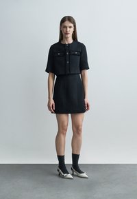 Žena stojí na jednoduchom pozadí, má na sebe čiernu krátku bundu s krátkymi rukávmi a zodpovedajúcu sukňu, čierne ponožky a biele lodičky.