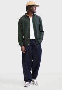 Homme portant une casquette beige, un cardigan boutonné vert foncé par-dessus une chemise blanche, un pantalon ample bleu marine, et des baskets argentées, debout les mains dans les poches.