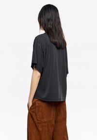 Camiseta negra de gran tamaño con mangas cortas, adornada con tachuelas decorativas en los hombros, combinada con pantalones de imitación ante marrones.