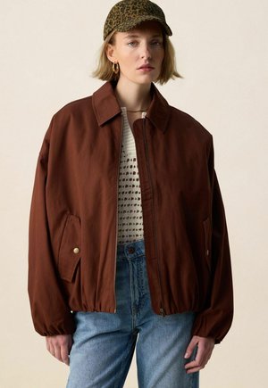 Femme portant une veste marron surdimensionnée, un haut blanc en maille, un jean bleu clair, une casquette à imprimé léopard et des boucles d'oreilles créoles dorées sur fond uni.