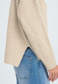Beige, geripptes Sweatshirt mit geometrischem Muster, seitlichen Schlitzen und langen Ärmeln, kombiniert mit blauer Denim. Strukturierter Strickstoff.