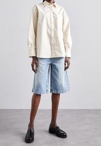 Camisa de manga larga color beige claro con cierre de botones, combinada con pantalones cortos de mezclilla azul hasta la rodilla. Mocasines negros completan el conjunto.