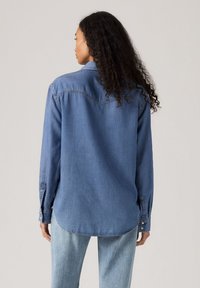 Denimskjorte i mellemblå med afslappet pasform, lange ærmer, knaplukning ved ærmerne og buet kant. Har stikningen detalje på ryggen.