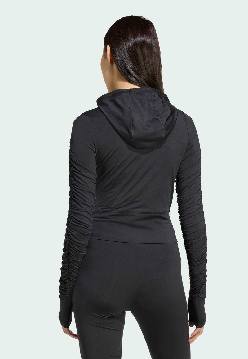 Donna che indossa un hoodie nero arricciato a maniche lunghe e leggings neri, vista di spalle su uno sfondo chiaro e semplice.