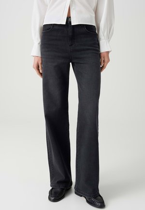 Person trägt schwarze Jeans mit weitem Bein, schwarze Schuhe und ein weißes langärmliges Hemd mit Manschetten und Knöpfen.