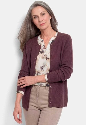 Femme aux longs cheveux gris portant un cardigan bordeaux boutonné sur une blouse à motifs et un pantalon beige, debout devant un fond uni.