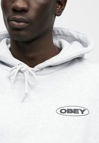 Ανοιχτό γκρι φούτερ με κουκούλα και κορδόνι, με το μαύρο λογότυπο "OBEY" σε οβάλ σχήμα στο στήθος. Μαλακή υφή υφάσματος με καθημερινό σχεδιασμό.