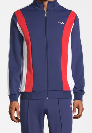 Mann trägt eine dunkelblaue FILA Trainingsjacke mit roten und weißen Seitenstreifen, Reißverschluss und passenden dunkelblauen Hosen.