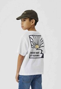 Camiseta blanca con un gráfico negro de un diseño de sol y ola; texto "HEAT WAVE // SURFING" debajo. Llevada con unos vaqueros azules y una gorra oscura.