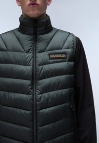 Vadderad väst i mörkgrön med hög krage och dragkedja. Har en quiltad design och en märkespatch på bröstet.