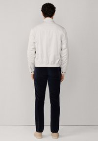 Hombre vistiendo una chaqueta de color claro, pantalones oscuros y zapatos beige, de pie frente a una pared gris claro y con las manos relajadas a los lados.