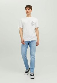 Camiseta blanca de algodón con mangas cortas y un estampado gráfico en el lado izquierdo del pecho, combinada con jeans de color azul claro y zapatillas negras.