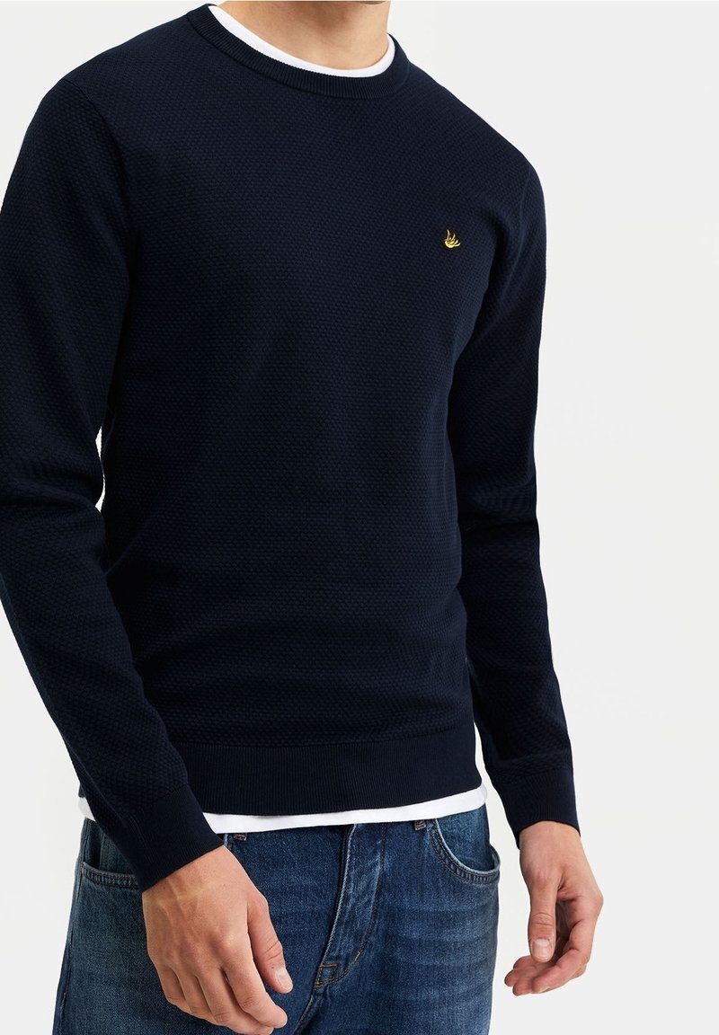 Marinefarbener strukturierten Pullover mit Rundhalsausschnitt, der ein kleines goldenes Logo auf der Brust und ein darunter liegendes weißes Shirt hat.