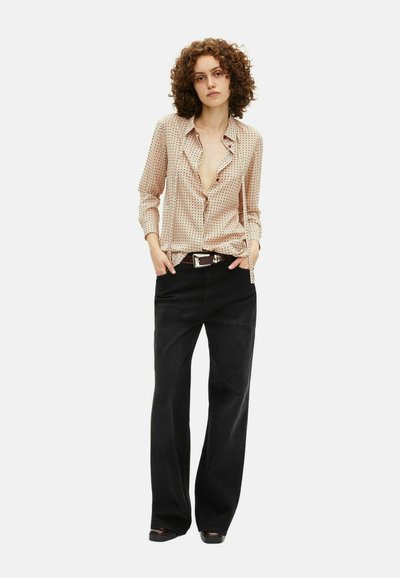 Donna con capelli ricci che indossa una blusa beige a pois, pantaloni neri a gamba larga, cintura marrone e scarpe marroni, in piedi con le mani nelle tasche.