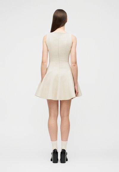 Γυναίκα που στέκεται με την πλάτη προς τα εμπρός, φορώντας ένα αμάνικο μπεζ φόρεμα με φούστα που φουσκώνει, άσπρες κάλτσες και μαύρα παπούτσια με τακούνι, σε απλό φόντο.