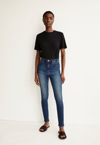 Schwarzes Kurzarm-T-Shirt, hoch geschnittene blaue Skinny-Jeans und dunkle Schlappen. Glattes Baumwollmaterial, klassisches Design, minimale Details.