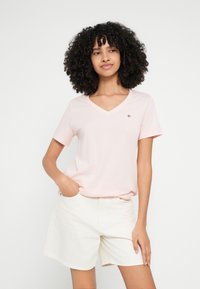 Világos rózsaszín, V-nyakú póló rövid ujjakkal, puha pamut anyagból, kis logóval a bal mellrészen, fehér rövidnadrággal párosítva.