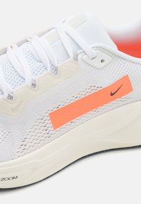 Bílé sportovní boty s síťovaným svrškem, s jasně oranžovým akcentem, texturovanými překryvy a polstrovanou podrážkou s černými detaily na podrážce.