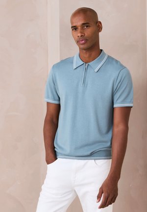 Muž v svetlomodrom tričku s krátkym rukávom a bielym lemom a bielych nohaviciach, stojaci pred jednoduchým béžovým pozadím, ruky vreckách.