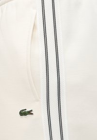 Λευκό ένδυμα με διαφανές πάνελ που διαθέτει δύο μαύρες κάθετες ρίγες και μικρό πράσινο λογότυπο κροκόδειλου κοντά σε πλαϊνή τσέπη.