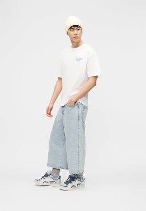 Noor mees, kes kannab valget mütsi, valget trükitud T-särki, laiu helesiniseid pükse, halli sokke ja valgeid-siniseid tenniseid, seisab ühevärvilise tausta ees.