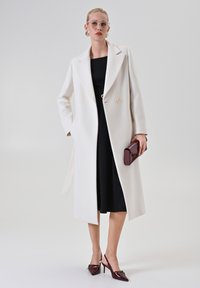 Donna che indossa un lungo cappotto bianco sopra a un vestito nero, tiene in mano una clutch bordeaux, con tacchi bordeaux abbinati e occhiali trasparenti.