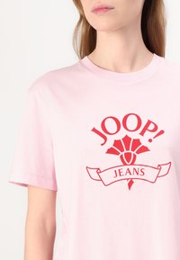 Světle růžové bavlněné tričko s červeným grafickým logem, obsahujícím nápis "JOOP!" a květinový emblém, doplněné bílou stuhou se slovem "JEANS."