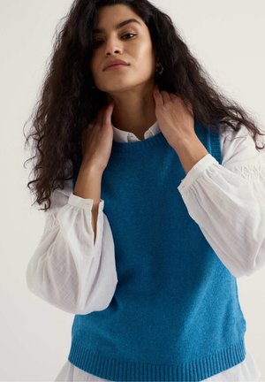 Mujer con cabello oscuro largo y rizado, vestida con un suéter sin mangas de punto azul sobre una blusa blanca de manga larga, ajustándose el cuello frente a un fondo liso.