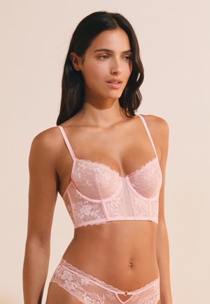 Frau mit langen dunklen Haaren, die ein rosa florales Spitzen-Dessous-Set trägt, blickt leicht seitlich vor einem schlichten beigen Hintergrund.